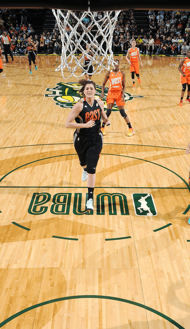 WNBA All-Star Game action: a player in an orange West team uniform goes up for a layup on the WNBA-branded court