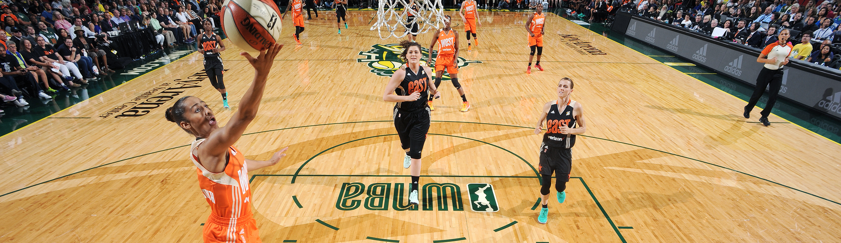 WNBA All-Star Game action: a player in an orange West team uniform goes up for a layup on the WNBA-branded court
